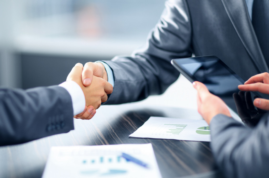 A picture of two people in suits shaking their hands over a desk,